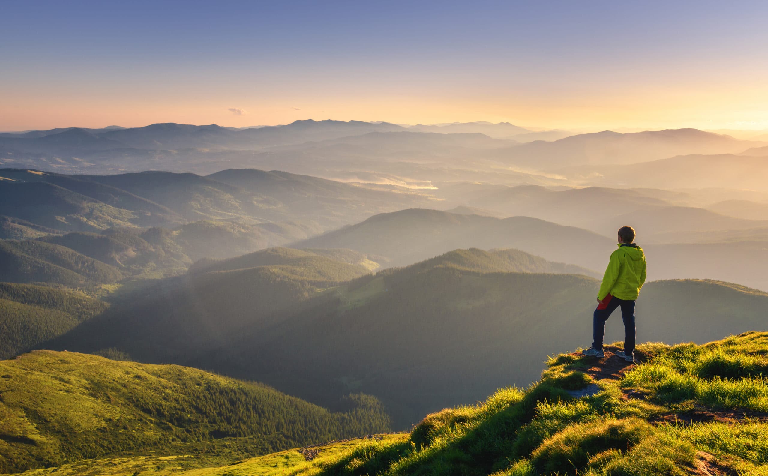 A male hiker stands at the top of a grassy ridge overlooking a mountain valley at sunset.