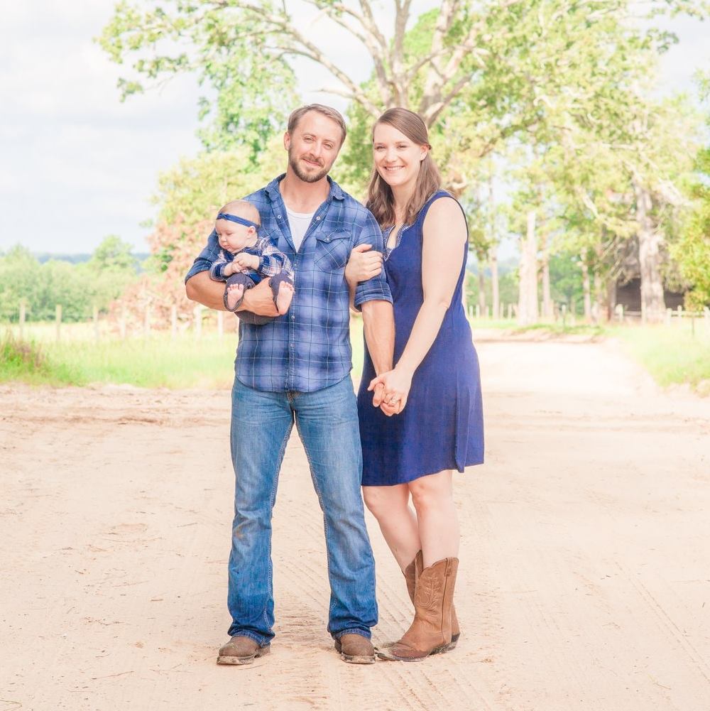 A man holding a baby stands next to a woman in the middle of a dirt road.